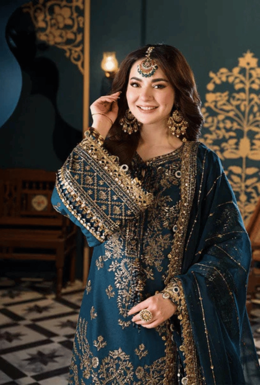 Woman in traditional blue and gold embroidered outfit with jewelry against a decorative wall.