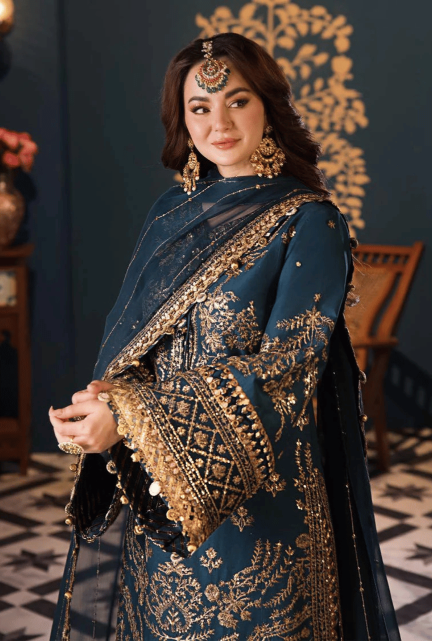 Woman wearing a blue and gold embroidered traditional outfit with jewelry against a dark background.