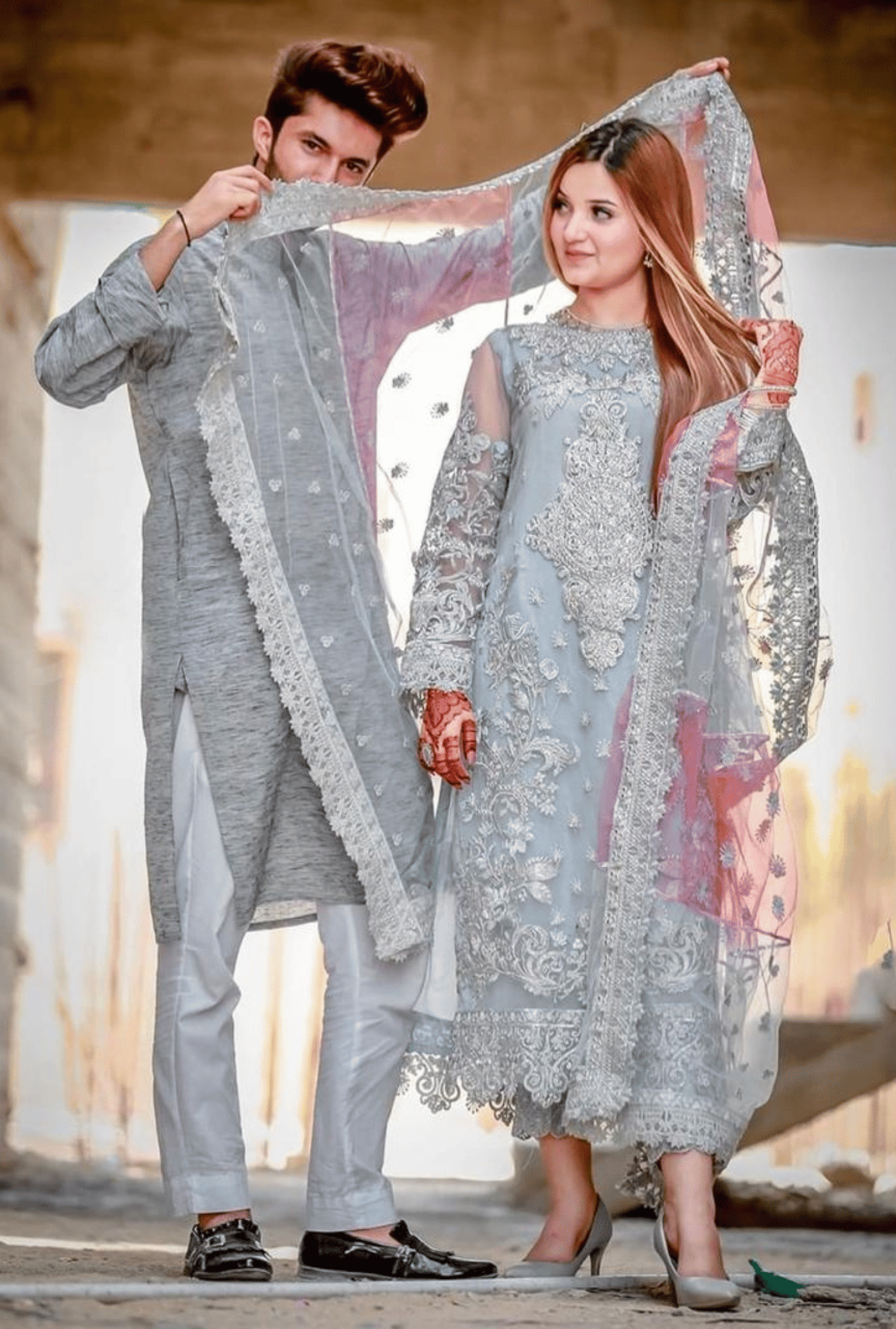 Man and woman in traditional light gray outfits with intricate designs, holding a sheer fabric.