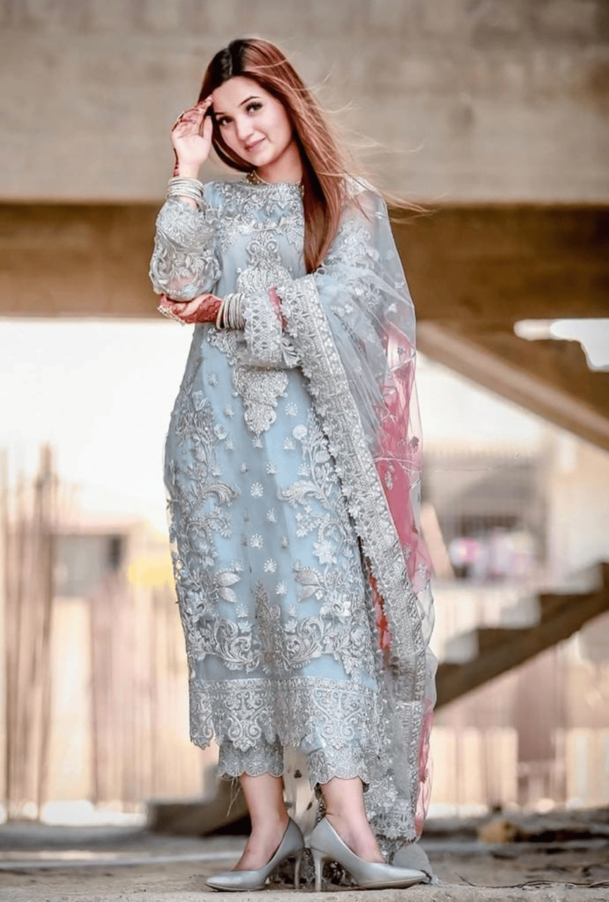 Woman in a light blue embroidered traditional outfit with a pink dupatta, standing outdoors.