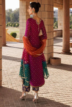 Woman in a traditional outfit with a pink and green dress and orange dupatta, standing in an outdoor setting.