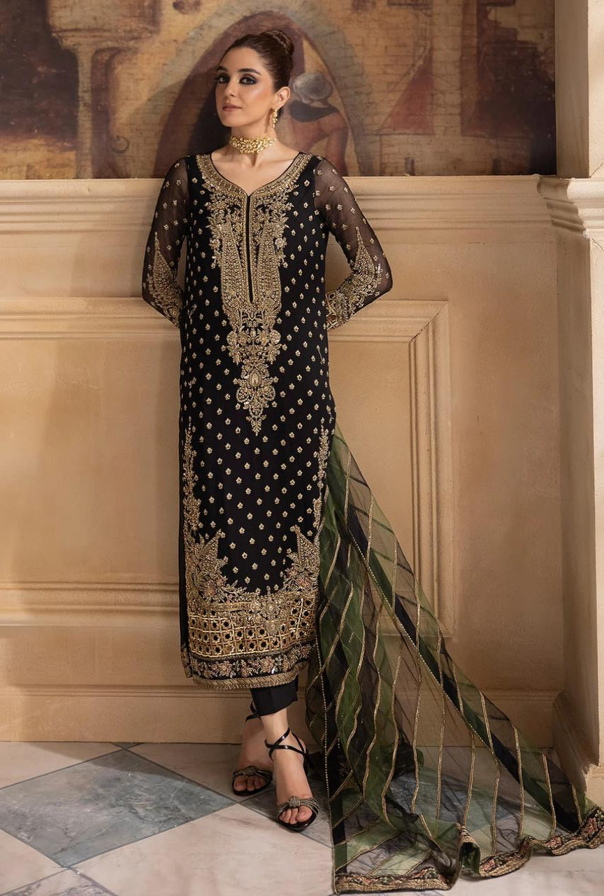 Woman in a black and gold embroidered traditional outfit with a green dupatta against a decorative wall.