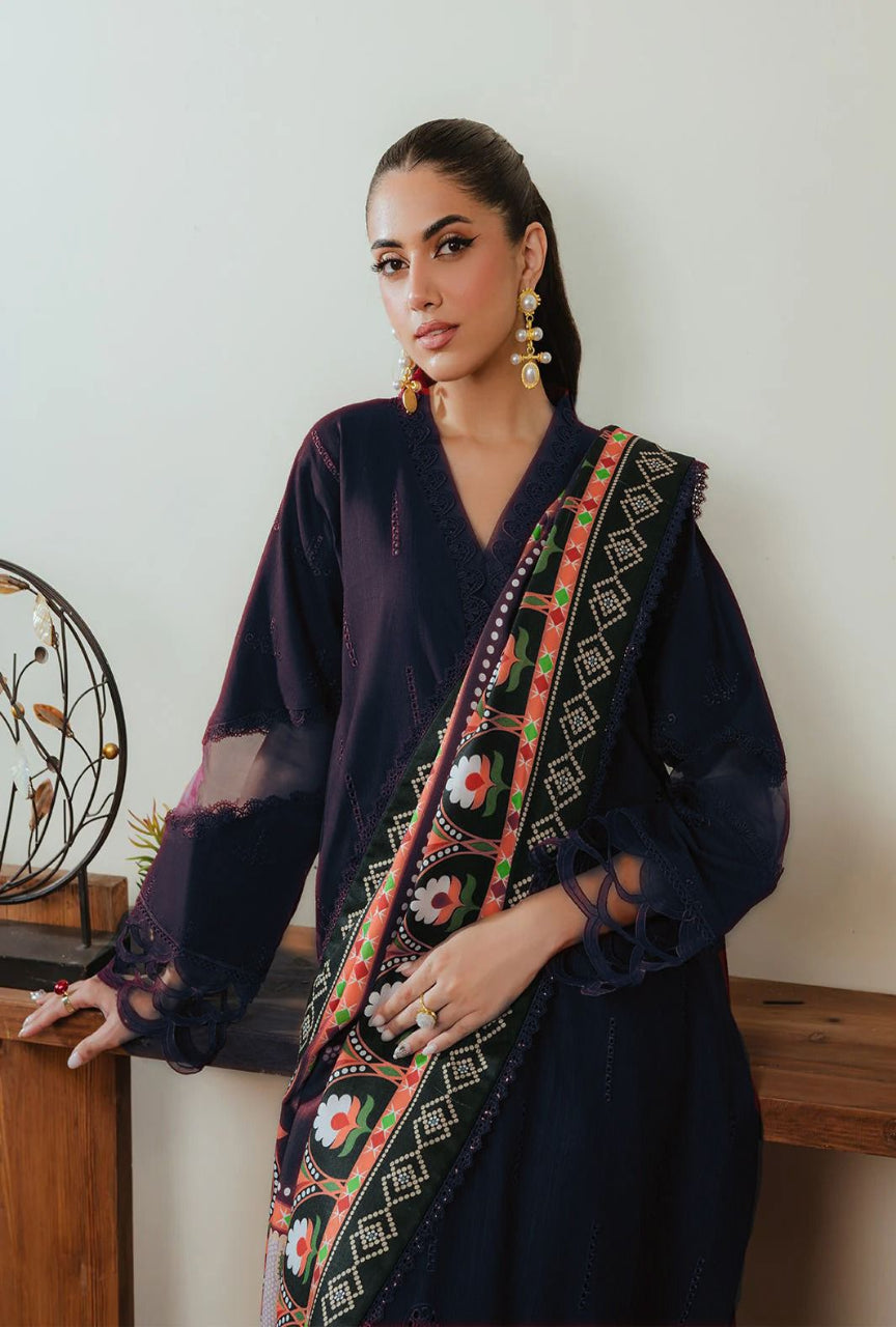 Woman wearing a dark blue traditional outfit with an embroidered multicolor scarf against a white wall.