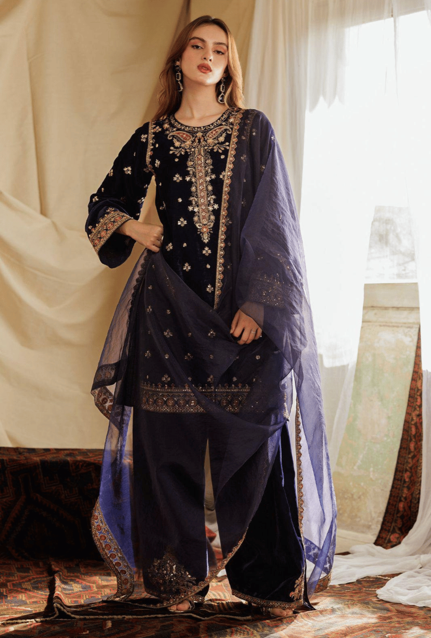 Woman wearing a dark blue embroidered traditional outfit with a sheer dupatta in a decorated indoor setting.