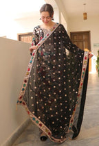 Woman holding a black embroidered saree with a colorful border indoors.