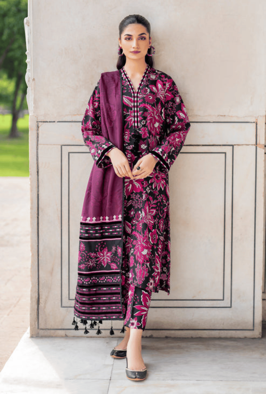 Woman wearing a traditional outfit with a purple dupatta and pink floral dress against a stone wall.
