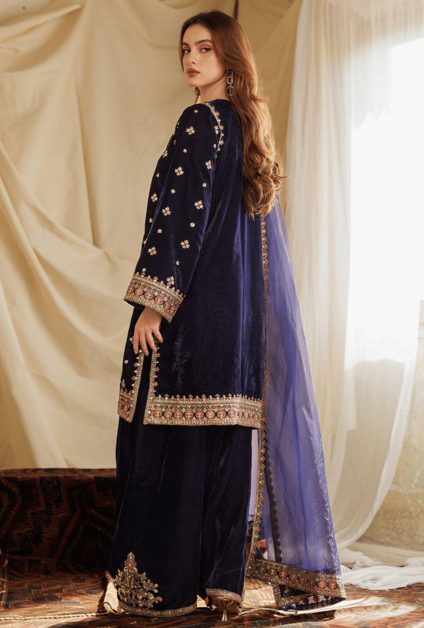 Woman in a navy blue embroidered outfit with a purple dupatta against a beige curtain background