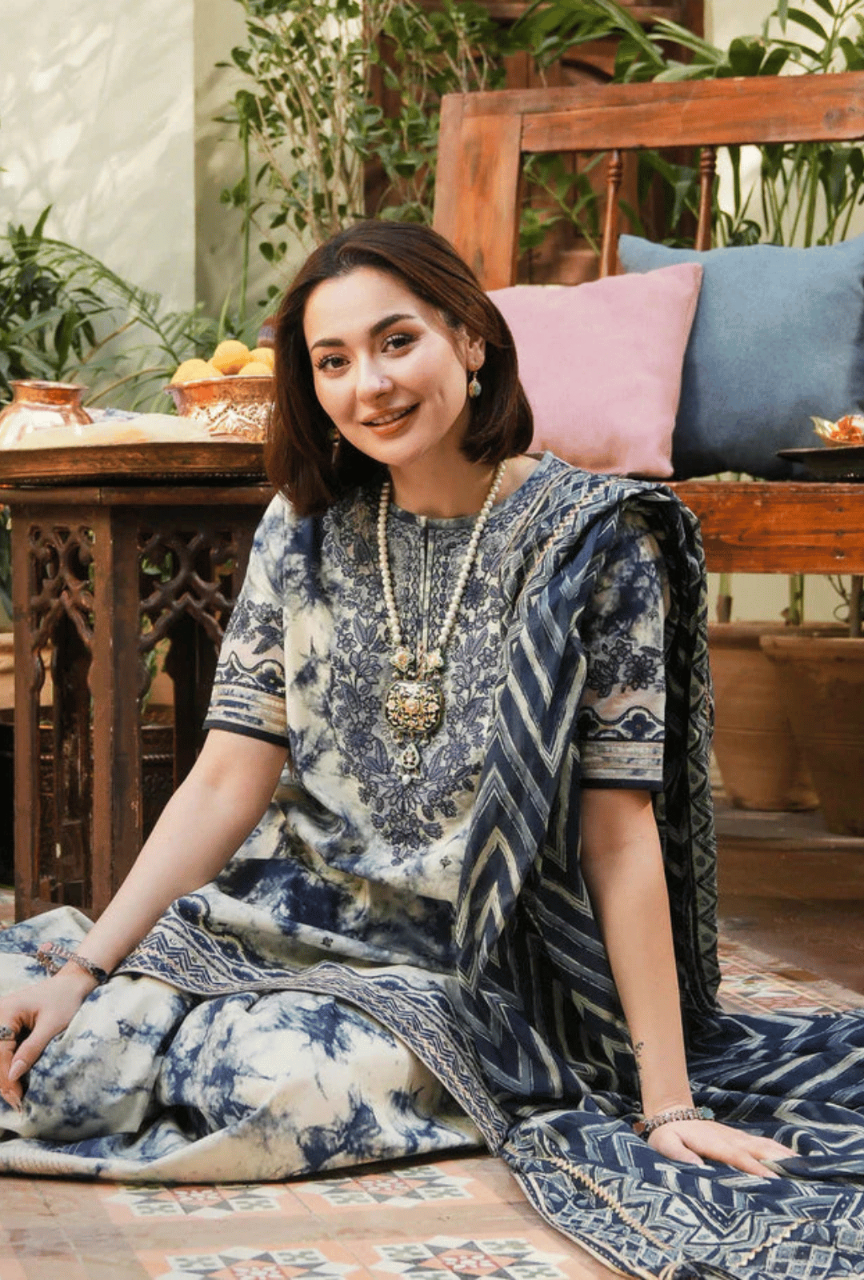 Woman in a blue and white patterned dress sitting on a wooden floor with plants and furniture in the background.
